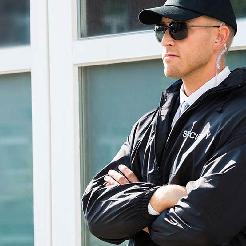 Agent de sécurité homme, uniforme noir, casquette, lunettes, oreillette. Bras croisés, "SECURITY" sur la veste. Vigilant.