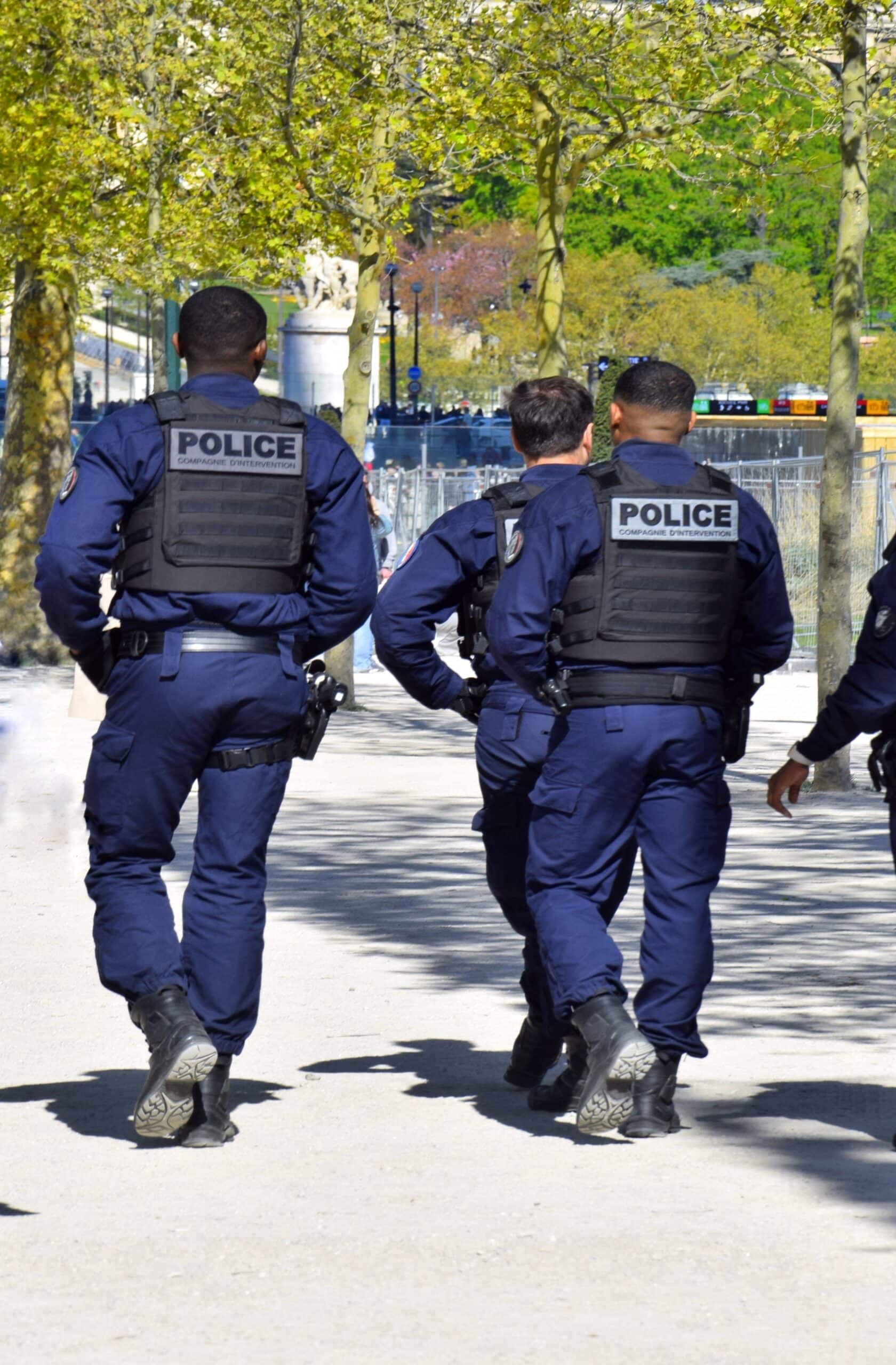 Trois policiers en uniforme bleu, gilets tactiques "POLICE COMPAGNIE D'INTERVENTION", marchent de dos sur un chemin arboré.