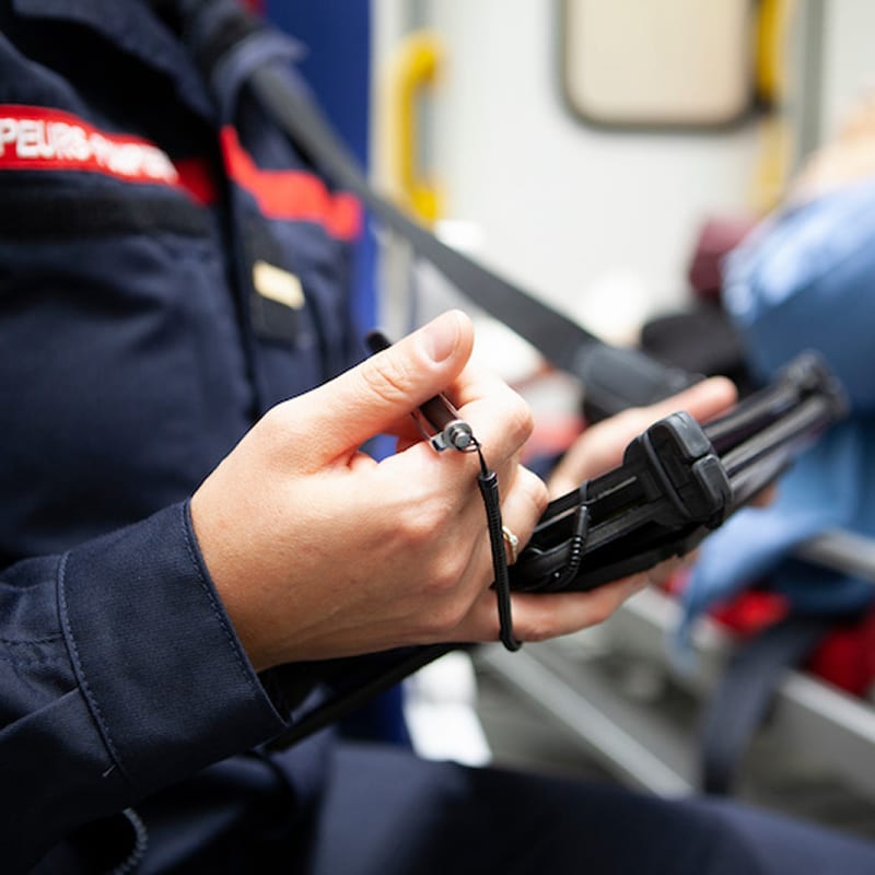 Sapeur-pompier en uniforme bleu et rouge, avec le texte SAPEURS-POMPIERS, utilise un stylet sur une tablette numérique, en contexte d'urgence.