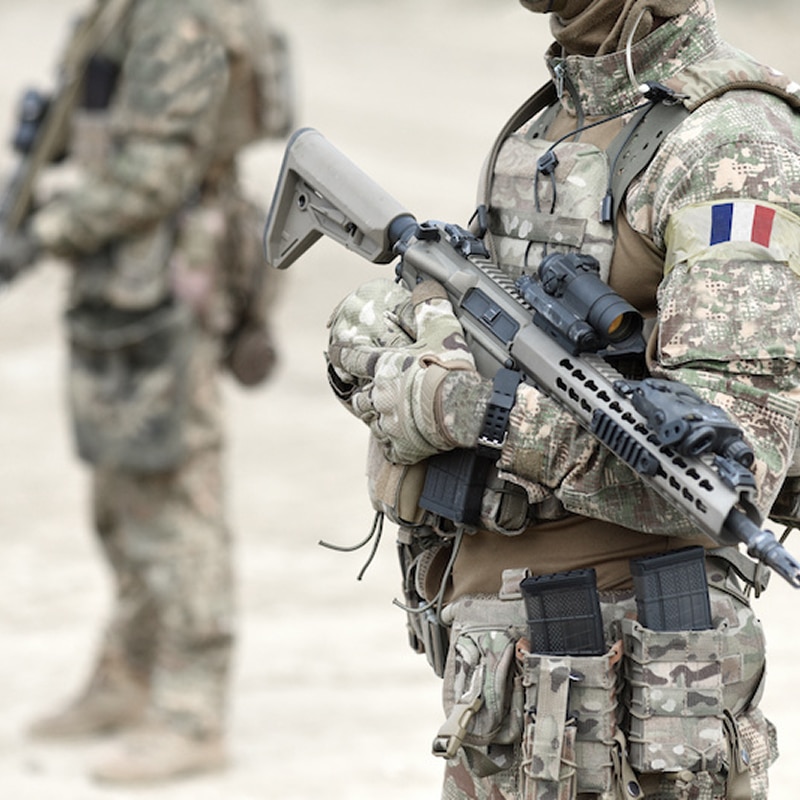 Soldat français en uniforme camouflage, fusil d'assaut à la main, avec un patch drapeau de la France. Un second soldat en arrière-plan.