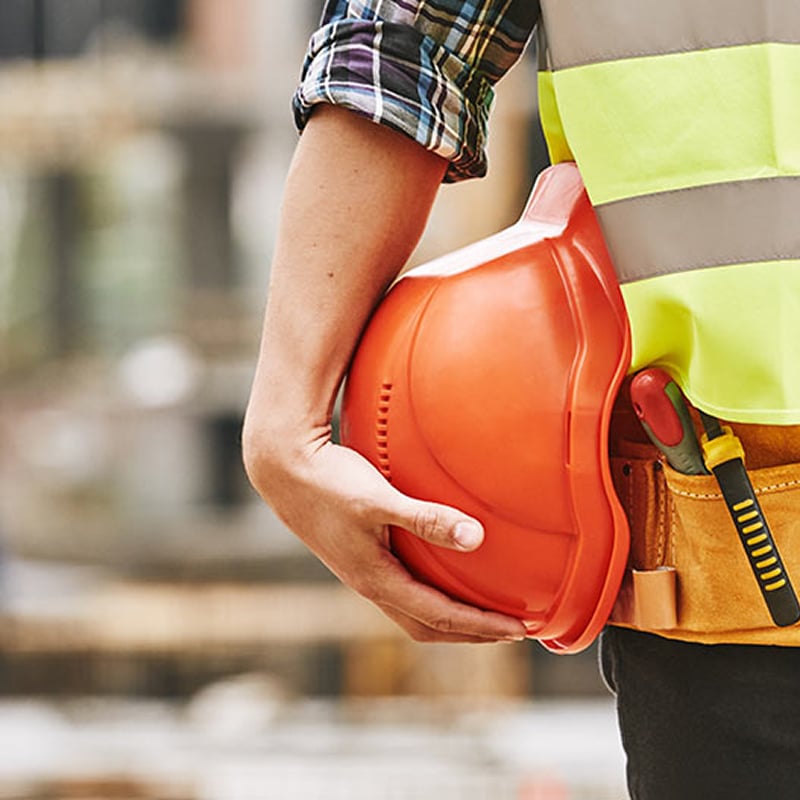 Travailleur en gilet fluo et chemise à carreaux tenant un casque de chantier orange, avec des outils à la ceinture.